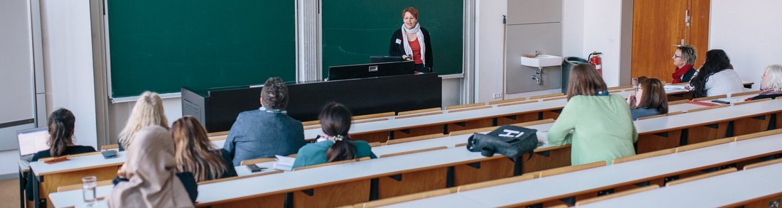 Mehrere Personen in einem Hörsaal, eine Person steht vorne, Präsentation sichtbar