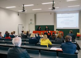 Mehrere Personen in einem Hörsaal, zwei Personen stehen vorne, Präsentation sichtbar