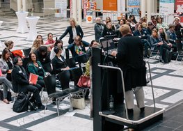 Blick auf das Publikum, eine Person steht im Publikum, eine Person steht vorne vor dem Mikrofon