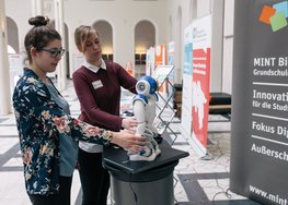 Zwei Frauen betrachten einen Roboter, Informationsstände im Hintergrund