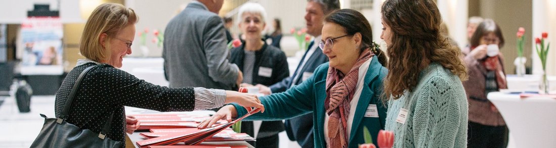 Drei Frauen stehen um einen Tisch, auf dem Veranstaltungsmappen liegen, weitere Personen im Hintergrund