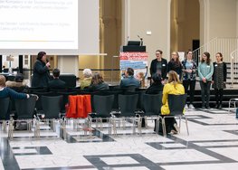 Blick von hinten, eine Frau und fünf Technikantinnen stehend, Publikum sitzend