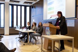 Professorin Barbara Schwarze stehend vor einem Mikrofon, im Hintergrund sitzen Technikantinnen