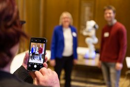 Eine Person macht mit ihrem Smartphone ein Foto, vor der Kamera stehen zwei Personen vor einem Roboterarm