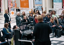 Blick auf das Publikum, eine Person steht im Publikum, eine Person steht vorne vor dem Mikrofon